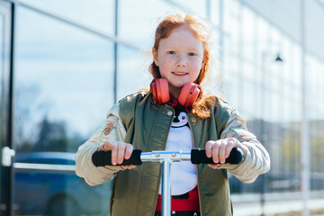 Closeup portrait of cute adorable smiling little curly red-haired caucasian girl child standing with scooter over business center on background outside, looking in camera, lifestyle childhood concept.