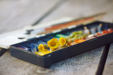 Wall Mural - On the wooden table is an old vintage palette with cuvettes, a set of brushes and a few blossoming flowers coltsfoot