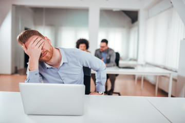 Upset businessman covering face with hand while sitting on front of laptop in coworking office.