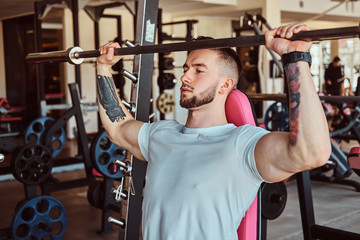 Young tattooed bodybuilder is doing his workout with barbell in gym. There are many sport equipment around.