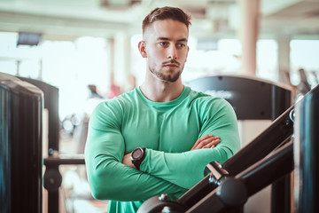 Young pensive bodybuilder in green shirt is standing near training apparatus with crossed hands.