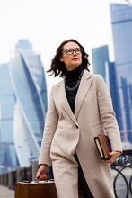 Beautiful Brunette In Light Coats With Books And Wooden Briefcase In Hand On The Background Of Skyscrapers