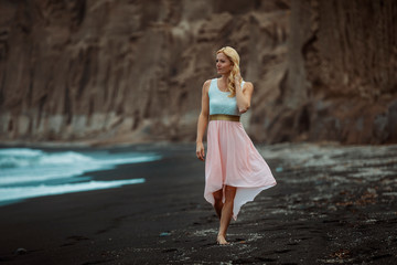 Wall Mural - blond woman on a black beach on Santorini, with cliff in the background and a rough sea