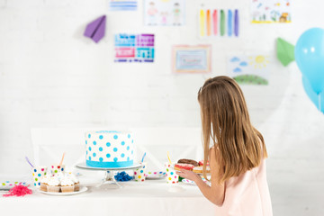 Wall Mural - back view of adorable kid holding plate with donuts at birthday table with cake during party