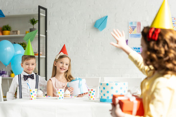 Wall Mural - kid waving to friends in party caps sitting at table during birthday celebration at home