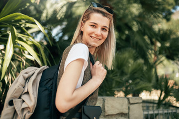 Portrait of young stylish woman walking on the street, wearing trendy outfit, with backpack on her shoulder, during her summer vacation. Young pretty traveler female posing on the street at sunny day.