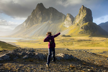 Happy joyful girl with long and dark hair jumping on nature in a outdoor hiking trip against the background of huge pointed, peaked mountains in eastern Iceland near the coast of the Atlantic Ocean.