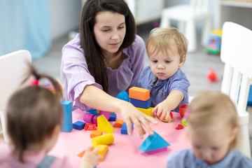 female teacher sitting at table in playroom with three kindergarten children constructing