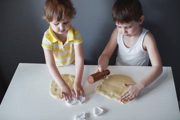 Little cheerful children make shortbread. Rolls out the dough with a rolling pin on a white table. The preparation of the dessert