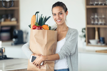 Beautiful young woman looking at camera while holding shopping bag with fresh vegetables in the kitchen.