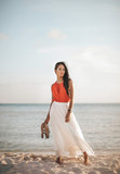 Fototapeta Na drzwi - gorgeous asian filipino girl posing in her 20s with an orange blouse and long white skirt in front of the ocean on the beach while holding her sandals on Turks and Caicos Islands looking at the camera
