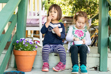 Two Young Girls Sitting On A Porch Together Eating A Snack.