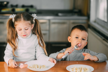 Wall Mural - Boy and girl children in the kitchen eating sausages with pasta is very fun and friendly