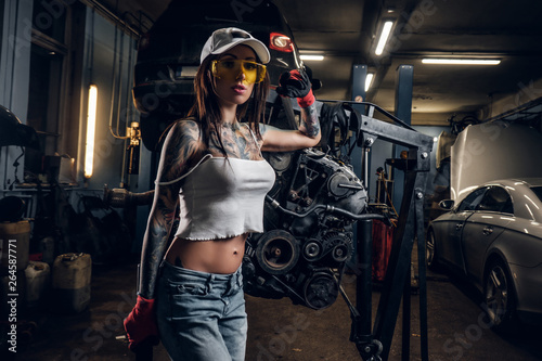 Fotografia  Sexual tattoed girl wearing cap and dirty clothes posing next to a car engine su