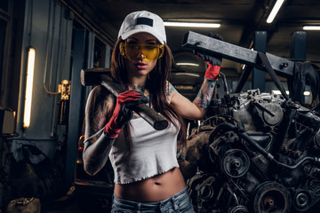 Stylish girl wearing cap and goggles posing with a steel hammer next to a car engine suspended on a hydraulic hoist in the workshop