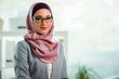 © Suzi Media  - Beautiful young working woman in hijab, suit and eyeglasses standing in office, smiling. Portrait of confident muslim businesswoman. Modern office with big window, man working at desk on background.