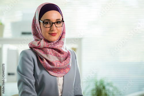 Fotografía Beautiful young working woman in hijab, suit and eyeglasses standing in office, smiling