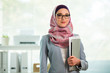 © Suzi Media  - Beautiful young working woman in hijab and eyeglasses smiling in office, holding folders and laptop in arms. Portrait of confident muslim businesswoman. Modern office with big window on background.