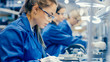 © Gorodenkoff - Female Electronics Factory Workers in Blue Work Coat and Protective Glasses Assembling Printed Circuit Boards for Smartphones with Tweezers. High Tech Factory with more Employees in the Background.
