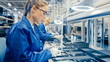 © Gorodenkoff - Woman Electronics Factory Worker in Blue Work Coat and Protective Glasses is Assembling Smartphones with Screwdriver. High Tech Factory Facility with more Employees in the Background.