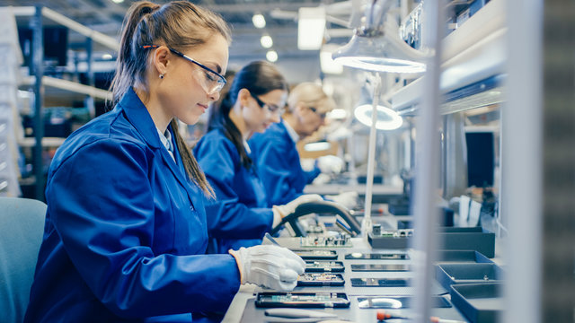 female electronics factory workers in blue work coat and protective glasses assembling printed circu