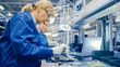 © Gorodenkoff - Female Electronics Factory Worker in Blue Work Coat and Protective Glasses is Assembling Smartphones with Screwdriver. High Tech Factory Facility with more Employees in the Background.