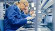 © Gorodenkoff - Female Electronics Factory Worker in Blue Work Coat and Protective Glasses is Assembling Smartphones with Screwdriver. High Tech Factory Facility with more Employees in the Background.