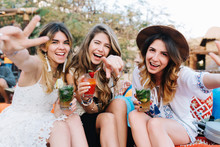 Attractive Long-haired Girls In Stylish Attires And Accessories Drink Cocktails And Posing With Hands Waving. Portrait Of Laughing Sisters Holding Glasses Of Soda, Having Fun On Picnic In Summer Day.