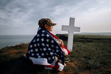 Child Teenager Wrapped In An American Flag Sits On The Grass Near The Grave Of His Father, A Soldier Hero Of America