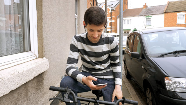 teen boy with smart phone listening or talking while sitting on bicycle british street. teenager and social media concept