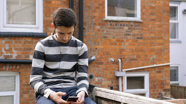 teen boy with smart phone listening or talking while sitting in british backyard garden. teenager and social media concept