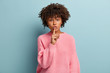 Do not slip word. Concerned black woman shushes at camera, keeps fore finger over lips, asks keep secret safe, dressed in loose pink jumper, isolated over blue background. Be quiet and mute.
