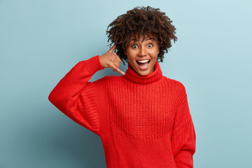 Happy overjoyed Afro American woman shows call me gesture, asks keep in touch, wears warm red sweater, being in high spirit, isolated over blue studio background. People and body language concept.