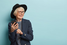 Old Attractive Woman Types Text Message On Cellular, Uses New Online Application, Has Positive Facial Expression, Looks Away, Wears Stylish Headgear And Coat, Stands Over Blue Studio Wall, Free Space