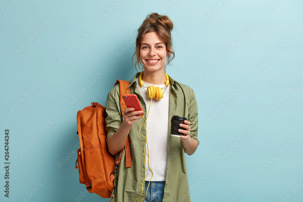 Studio shot of glad female student has coffee break after lectures, listens audio book in headphones, enjoys record from website, uses mobile phone for chatting online, has rucksack on back. - obrazy, fototapety, plakaty 