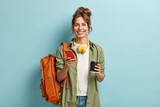 Fototapeta  - Studio shot of glad female student has coffee break after lectures, listens audio book in headphones, enjoys record from website, uses mobile phone for chatting online, has rucksack on back.