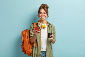 studio shot of glad female student has coffee break after lectures, listens audio book in headphones
