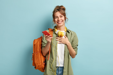 Indoor shot of pleased European student happy to receive message of congratulation after passing exam, holds palm on chest, demonstrates gratitude, listens music with headphones, wears stylish clothes