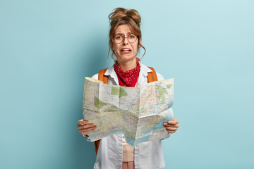 crying female tourist lost in unknown destination, holds map, tries to find way, looks with dissatis