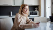 © Ulia Koltyrina - beautiful young woman in a cozy sweater sits in the kitchen and uses wireless headphones for smartphone.