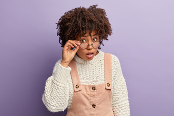 Wall Mural - Photo of curious dark skinned lady looks surprisingly curiously through round glasses, can not believe in shocking news, wears oversized white jumper and overalls, stands over purple background