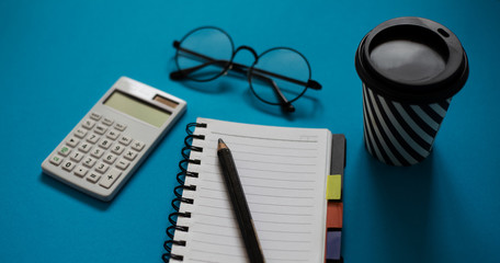 Top view of blue office desk, with cup of coffee, empty notebook and pencil, glasses and white calculator.