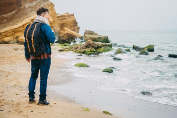Wall Mural - traveler with a backpack standing near a rock against a beautiful sea with waves, a stylish hipster 