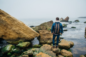 Wall Mural - traveler with a backpack stands on a rock against a beautiful sea with waves, a stylish hipster boy 