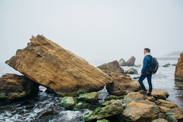 Wall Mural - Traveler with a backpack stands on a rock against a beautiful sea with waves, a stylish hipster boy posing near a calm ocean during a wonderful journey around the world. Shoot from the back