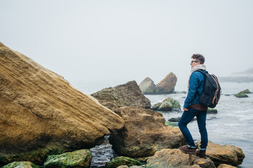 Wall Mural - traveler with a backpack stands on a rock against a beautiful sea with waves, a stylish hipster boy 