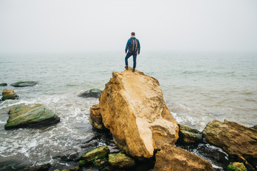Wall Mural - traveler with a backpack stands on a rock against a beautiful sea with waves, a stylish hipster boy 