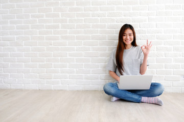 Young asian woman showing ok hand sign and smiling while working with laptop computer in white room with copy spce, people with positive gesture