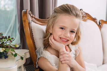 Portrait of a little attractive smiling girl in a white dress with curled hair against the background of a grunge wall.