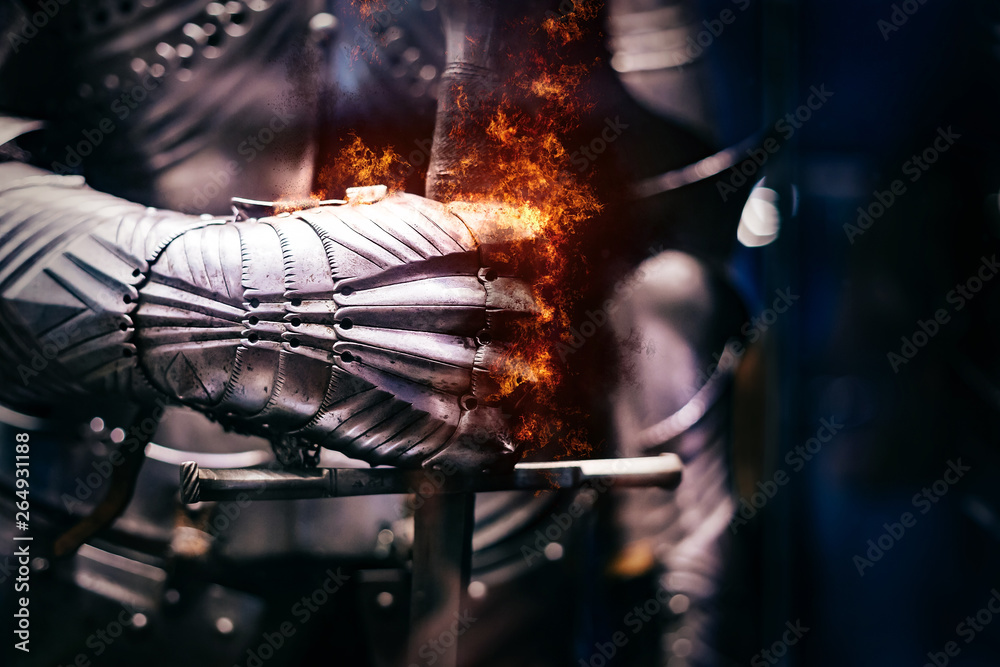 Close up of a Medieval steel armour with iron glove hand bursting with flames of fire, holding a giant sword - obrazy, fototapety, plakaty 